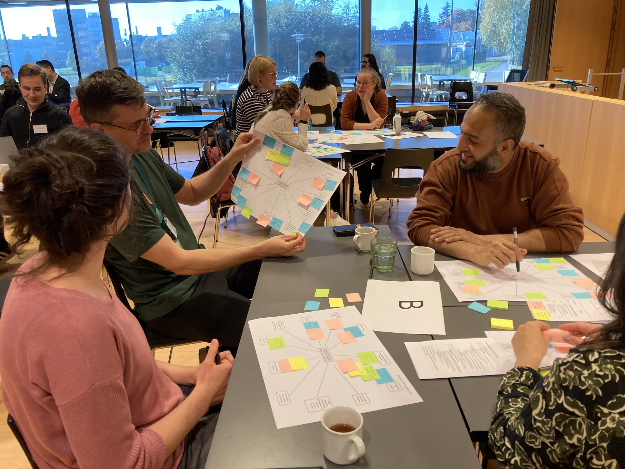 Participants work in small groups during a workshop, using sticky notes and visual templates. The groups discuss and organize their ideas around a shared task.